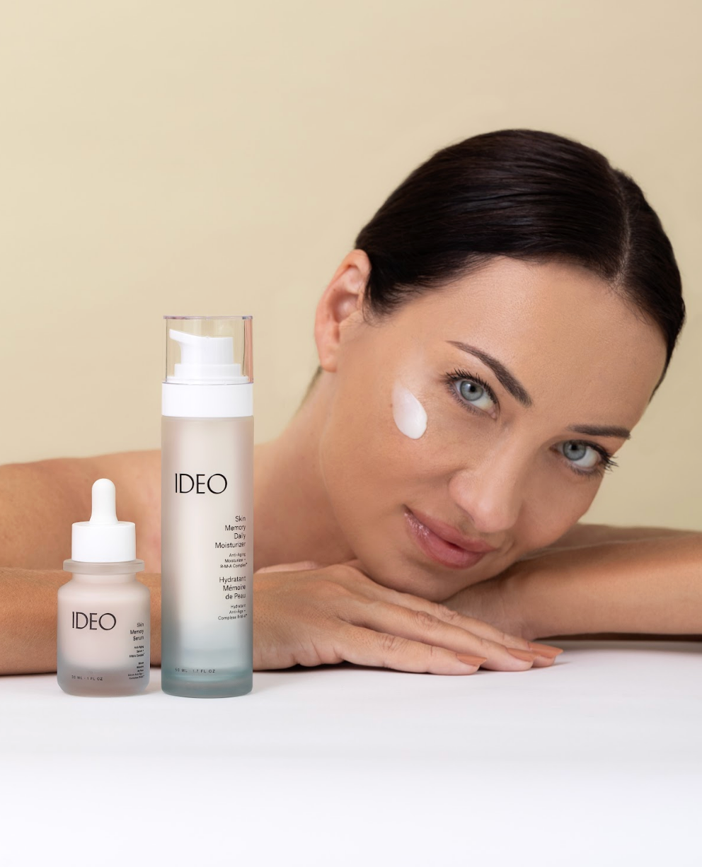 Woman resting chin on hands with IDEO Skin Memory Moisturizer applied to cheek beside Skin Memory Serum and Moisturizer bottles on white surface lifestyle portrait